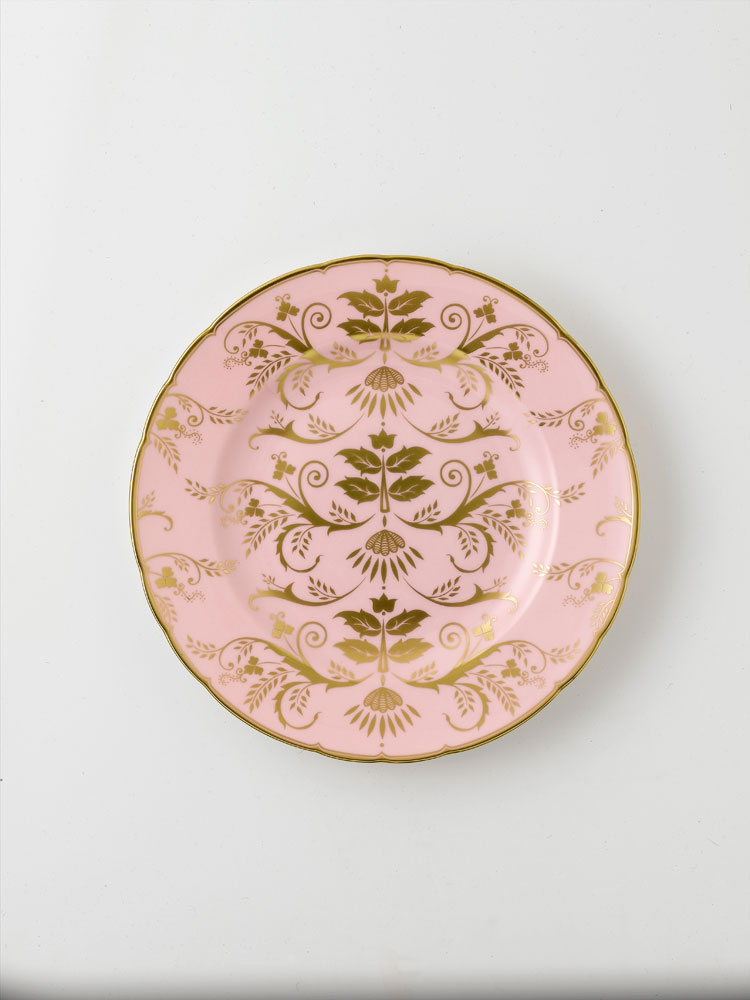 A round pink salad plate with gold floral patterns and a gold rim.
