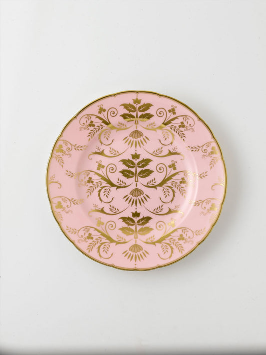 A round pink salad plate with gold floral patterns and a gold rim.
